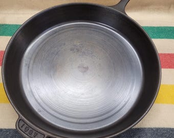 May include: A round black cast iron skillet with a smooth, polished interior surface showing concentric circular markings. The pan features a long handle and a smaller helper handle with the embossed text AB&I visible on the helper handle.