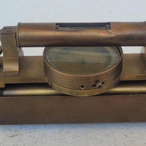 May include: A vintage brass surveying instrument featuring a central circular compass housing, a horizontal spirit level tube mounted on top, two adjustment knobs on either side, and a cylindrical telescope extending from both ends of the base.
