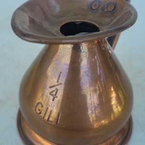 May include: A copper measuring vessel with a wide, flared rim and a single handle. The body features an engraved inscription that reads 1/4 GILL, indicating its capacity. The metal surface displays a warm, metallic sheen with subtle color variations.