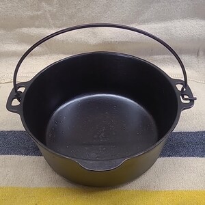 May include: A round black cast iron pot featuring a smooth interior surface, a wide open top, and two side handles with integrated loops. A thin, arched metal bail handle is attached to the side loops, extending upward over the open pot.