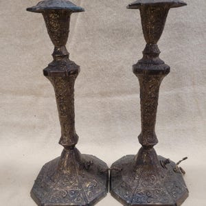 May include: A pair of dark bronze-toned metal candlesticks featuring intricate embossed scrollwork patterns, hexagonal bases, and flared tops. Each candlestick includes a small electrical component at the base and a wire extending from the side.