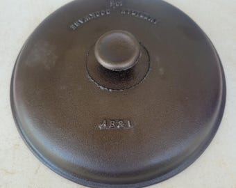 May include: A round, dark brown cast iron lid featuring a central knob handle. The surface is embossed with the text AB&I and AMERICAN COOKWARE along with the number 10. The lid has a smooth, matte finish and a slightly raised outer rim edge.