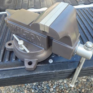 May include: A grey Wilton bench vise with a swivel base and a silver handle. The side of the tool features the embossed text WILTON SCHILLER PARK ILL USA. The vise includes a flat anvil surface and a sliding jaw mechanism with a metallic finish.