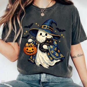 May include: A charcoal grey short-sleeve t-shirt featuring a graphic of a white ghost wearing a blue witch hat and cape, holding a jack-o'-lantern bucket filled with sweets and a black cat. The design includes small yellow stars and black bats.