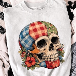 Peut inclure: Un sweat-shirt blanc à col rond arborant un motif central représentant un crâne humain composé de divers empiècements en tissu aux motifs vichy et floraux rouges, bleus et verts, entouré d'un bouquet de fleurs d'hibiscus rouges et orange.