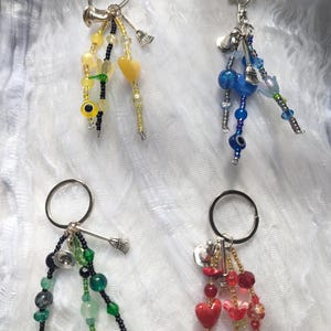 May include: Four keychains featuring metal rings and dangling beaded strands. One is yellow with heart and eye beads, one blue with a star charm, one green with black accents, and one red with heart-shaped beads. Each includes small silver-toned charms.