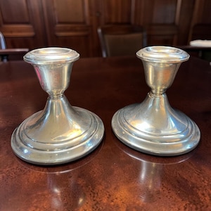 May include: A pair of silver-toned metal candle holders featuring a wide, circular base, a tapered stem, and a flared top rim designed to securely hold a single candle. The metallic surface has a polished finish with subtle reflective highlights.