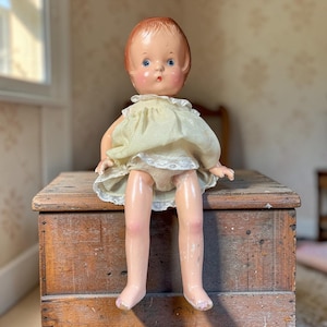 May include: A vintage doll with molded reddish-brown hair, blue eyes, and rosy cheeks. The doll wears a pale yellow sleeveless dress featuring a ruffled collar and white lace trim along the hemline, with visible paint wear on the legs and feet.