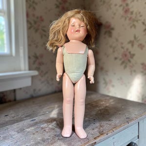 May include: A doll with light brown hair, a pale pink face, and closed eyes. The doll features a light green fabric torso piece and smooth, light-colored limbs. The doll stands upright with arms positioned at the sides and a neutral facial expression.