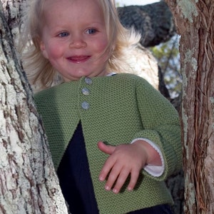 May include: A young girl with blonde hair is wearing a green knitted cardigan with buttons and a dark blue skirt. She is standing next to a tree trunk.