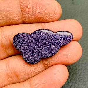 May include: A cloud-shaped cabochon made of dark blue goldstone, featuring a smooth, polished surface with embedded metallic glitter particles that create a sparkling, starry effect across the deep purple-blue material.