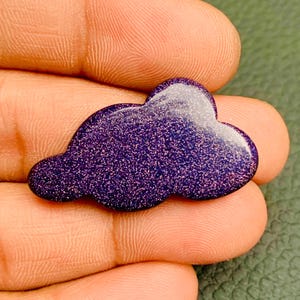 May include: A cloud-shaped cabochon made of dark blue goldstone, featuring a smooth, polished surface with embedded glittery particles that create a sparkling effect. The piece has a rounded, organic outline with multiple lobes.