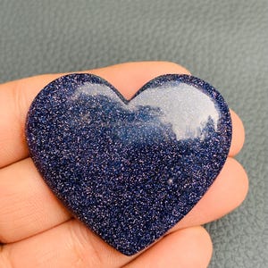 May include: A heart-shaped blue sandstone piece with a smooth, polished surface. The material is a deep, dark blue color filled with numerous tiny, reflective, sparkling metallic flecks that create a starry, glittery appearance across the entire object.