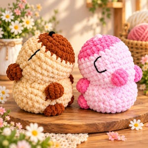 May include: Two plush crochet toys shaped like rounded creatures. The left toy is cream and brown with closed eyes. The right toy is pink and light pink with closed eyes. Both have textured yarn surfaces and small rounded limbs.