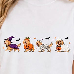 May include: A white t-shirt featuring a graphic of four golden retriever dogs dressed in Halloween costumes. The dogs are depicted as a witch, a pumpkin, a mummy, and a ghost holding a jack-o'-lantern bucket, accented by small bats and stars.