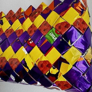 May include: A handbag constructed from woven strips of colorful snack food packaging. The surface features a repeating diagonal pattern of bright yellow, deep purple, and red graphic elements, including visible text fragments like arbol.
