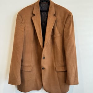 May include: A tan blazer with a soft, suede-like texture, featuring a notched lapel, two dark buttons on the front, two flap pockets, and a chest pocket. The interior is lined with a dark fabric, and the garment is displayed on a black hanger.