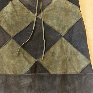 Op de afbeelding: Een suède rok met een ruitvormig patchworkpatroon in donkerbruin en olijfgroen. Het kledingstuk is voorzien van een taille met trekkoord en dunne suède veters in het midden, opgebouwd uit afwisselende donkerbruine en olijfgroene suède panelen.