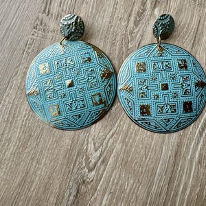 May include: A pair of round, light blue earrings featuring a detailed, embossed geometric pattern with metallic gold accents. Each earring has a smaller, matching circular stud top connected by a small metal ring to the larger circular pendant.