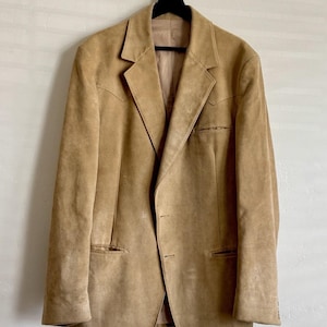 May include: A tan suede blazer featuring a notched lapel, two front buttons, and western-style pointed yokes on the chest. The jacket includes two lower welt pockets and one chest welt pocket, finished with a smooth interior lining.