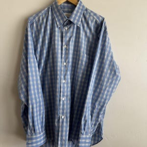 May include: A long-sleeve button-down shirt featuring a blue and white plaid pattern. The shirt has a standard collar, a front button placket with white buttons, and buttoned cuffs. The fabric displays a grid-like check design throughout.
