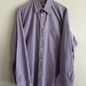 May include: A long-sleeved button-down shirt featuring a vertical striped pattern in shades of purple, white, and thin orange lines. The shirt includes a pointed collar, a front button placket, a single chest pocket, and buttoned cuffs on the sleeves.