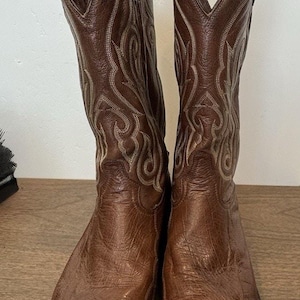 May include: A pair of brown leather cowboy boots featuring a textured ostrich-skin vamp, a pointed toe, and decorative light-colored stitching patterns on the tall shafts. The boots include pull straps at the top and a stacked heel design.
