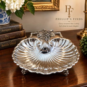 May include: A silver-toned metal serving dish shaped like a scallop shell, featuring a decorative grape cluster and leaf motif at the top edge. The dish has a ribbed surface and stands on three small feet, each adorned with a tiny cluster of grapes.