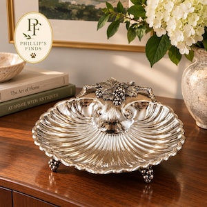 May include: A silver-toned metal serving dish shaped like a large scallop shell. The dish features intricate embossed grape clusters and vine leaves at the top center and rests on three small feet, each decorated with a cluster of grapes.