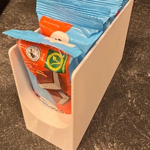 Könnte beinhalten: Ein weißer Kunststoff-Aufbewahrungsbehälter mit vier hellblauen Snack-Packungen. Jede Packung hat einen orangefarbenen Akzentstreifen, eine weiße kreisförmige Grafik und ein grünes Vegetarisch-Siegel. Die Packungen stehen in einer Reihe im Behälter.