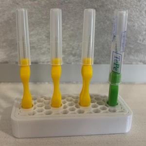 Könnte beinhalten: Ein weißer, rechteckiger Ständer mit Wabenmuster hält drei gelbe Interdentalbürsten und eine grüne TePe Interdentalbürste. Jede Bürste ist mit einer transparenten Kunststoff-Schutzkappe über den Borsten versehen.