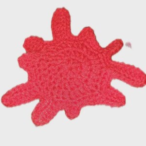 May include: A bright red crochet appliqué shaped like an irregular paint splatter or ink blot. The piece features a textured, looped yarn construction with multiple pointed extensions radiating from a central, rounded core, creating a distinct shape.