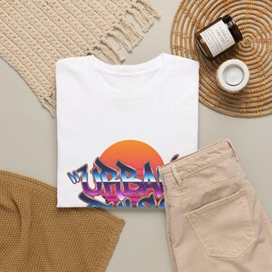 Peut inclure: Un t-shirt blanc à manches courtes avec une impression graphique d'un soleil orange vif se couchant derrière un lettrage de style graffiti indiquant URBAN PULSE dans des tons de bleu, violet et rose avec un motif de grille horizontale.