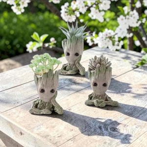 May include: Three light brown, tree-like humanoid planters with textured bark surfaces. Each planter features a unique pose and holds a small green succulent or air plant. The figures have large, dark, oval eyes and a smooth, smiling facial expression.