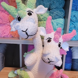 May include: A white crocheted two-headed dragon with green and pink horns. The dragon is sitting on a wooden surface.