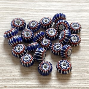 Può includere: Una collezione di perline di vetro blu, rosse e bianche. Le perline hanno un design floreale con un foro centrale. Le perline sono rotonde e cilindriche, con un motivo a strisce. Le perline sono sparse su una superficie chiara.