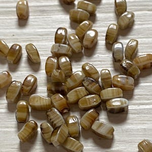 Pode incluir: Uma coleção de pequenas contas retangulares com bordas arredondadas, apresentando um acabamento brilhante e um padrão marmorizado em tons de castanho, marrom e creme. Cada conta tem uma superfície lisa e polida com faixas horizontais visíveis.