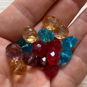 May include: A collection of small, faceted, translucent beads in vibrant shades of red, blue, and amber. Each bead features a multi-faceted surface that catches the light, creating a sparkling effect. The beads are round and have a central hole.
