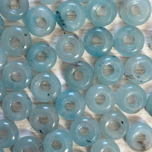 Könnte beinhalten: Eine Sammlung von hellblauen donutförmigen Perlen mit einer glatten, polierten Oberfläche. Die Perlen haben ein Loch in der Mitte und sind aus Naturstein.