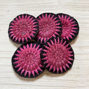 May include: Five pink and black embroidered round patches with a circular design. The patches are made of fabric and have a stitched edge.