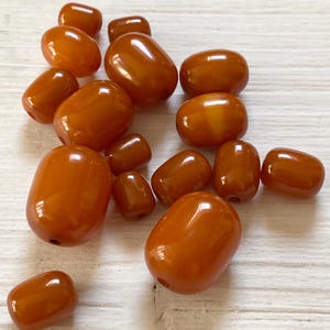 Könnte beinhalten: Eine Ansammlung glatter, fassförmiger Perlen in einer durchscheinenden, warmen bernstein-orangen Farbe. Jede Perle hat eine polierte, glänzende Oberfläche und ein Mittelloch zum Auffädeln, variiert in der Größe und liegt verstreut.