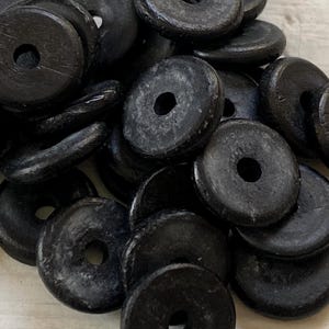 Può includere: Una collezione di perline circolari in legno nero, ciascuna dotata di un foro centrale. Le perline presentano una finitura opaca e testurizzata e sono ammassate insieme, mostrando variazioni nella venatura del legno e nell'usura superficiale.