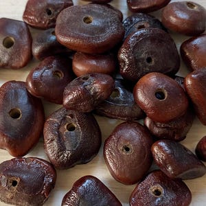 May include: A collection of dark brown, irregularly shaped, flattened organic beads. Each bead features a smooth, polished surface with a visible central hole, showcasing natural variations in texture and deep mahogany tones throughout the group.