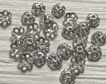 May include: A group of silver metal bead caps with a decorative openwork design. The caps are scattered on a white wooden surface.