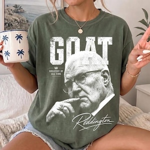 May include: An olive green t-shirt featuring a large white graphic of a man wearing glasses with the text GOAT, GREATEST OF ALL TIME, and Reddington in a script font. The shirt has a crew neckline and short sleeves, displaying a vintage-style print.