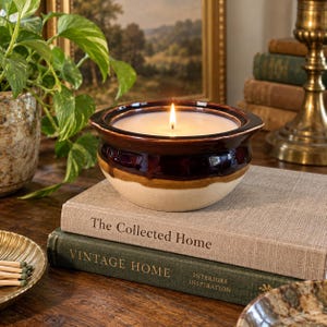 May include: A lit candle in a two-toned ceramic bowl with a dark brown glazed rim and a cream-colored base. The candle features a single burning wick centered in white wax, resting on a stack of two hardcover books titled The Collected Home.