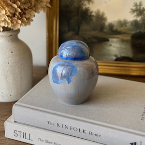 May include: A small, rounded ceramic jar with a lid, featuring a glossy, light gray base glaze accented by vibrant, crystalline blue floral-like patterns on the body and the lid. The jar has a smooth, reflective surface and a tapered neck.