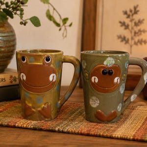 May include: Two ceramic mugs featuring raised, sculpted animal faces. The left mug is mustard yellow with blue polka dots and a brown frog-like face. The right mug is sage green with white polka dots and a brown monkey-like face, both with handles.