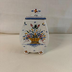 May include: A white ceramic jar with a lid, featuring a hand-painted yellow basket filled with blue, red, and green floral motifs. The jar has a tapered shape, stands on three small feet, and includes a matching lid with a decorative finial.