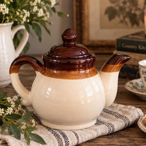 Puede incluir: Una tetera de cerámica con cuerpo de color crema y tapa, asa y boquilla esmaltadas en marrón brillante. La tetera tiene una forma redondeada con una banda marrón distintiva alrededor de la parte superior y un pomo marrón a juego en la tapa.